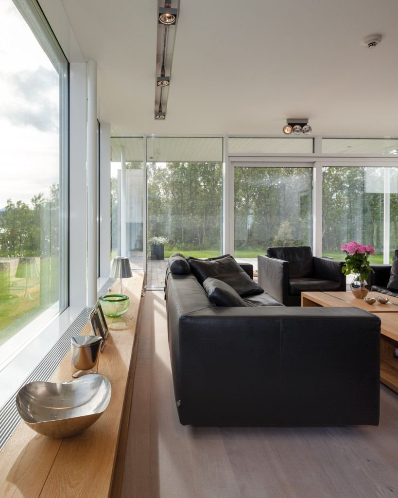 Stue i Villa Sirilund med sorte skinnsofaer foran vindusfelt fra gulv til tak og trær utenfor.